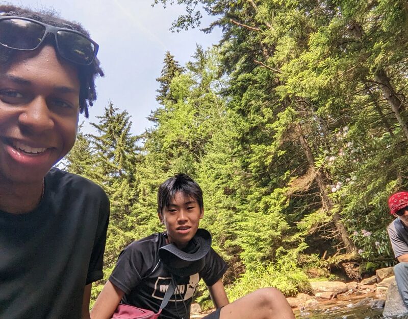 The image shows three young people in a natural setting, possibly a forest or park. Two of them are prominently featured in the foreground, with one wearing sunglasses and a black t-shirt, and the other sitting on a rock near a stream. The third person is partially visible in the background. The environment includes lush greenery and a flowing stream, suggesting an outdoor adventure or leisurely activity.